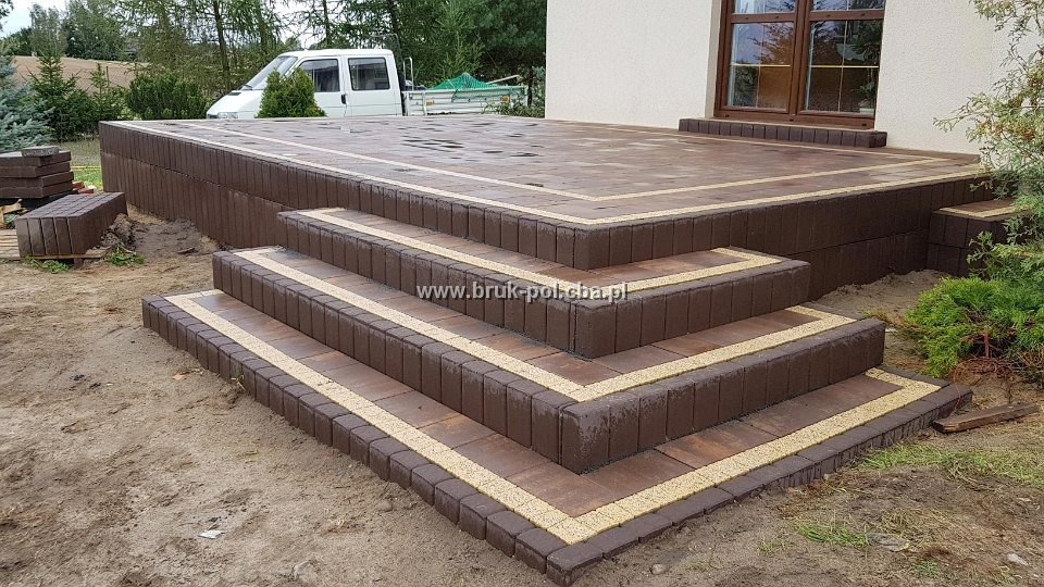 Schody wejściowe i taras wyłożone brązową kostką brukową z jasnymi akcentami, widoczny samochód dostawczy w tle.