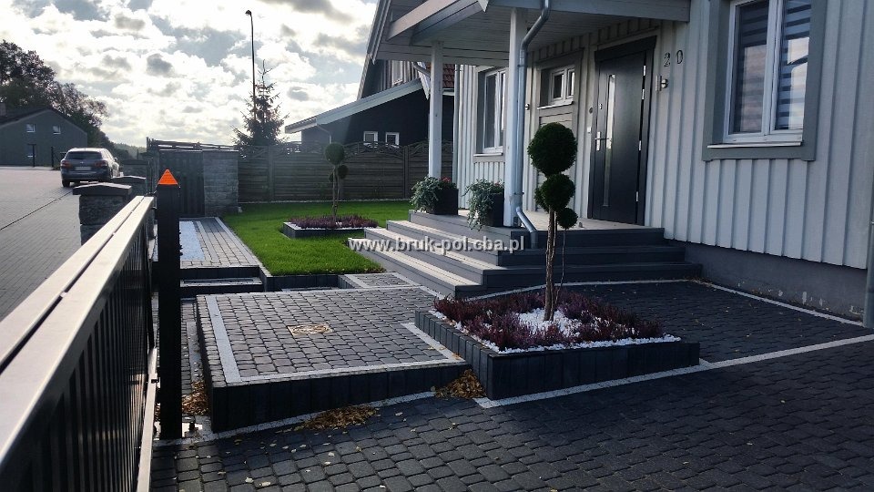 Ułożona, ciemnoszara kostka brukowa przed domem z szarymi schodami i zadbanym trawnikiem, obrzeża z białych kamyków, widoczne drzewka ozdobne i brama wjazdowa.