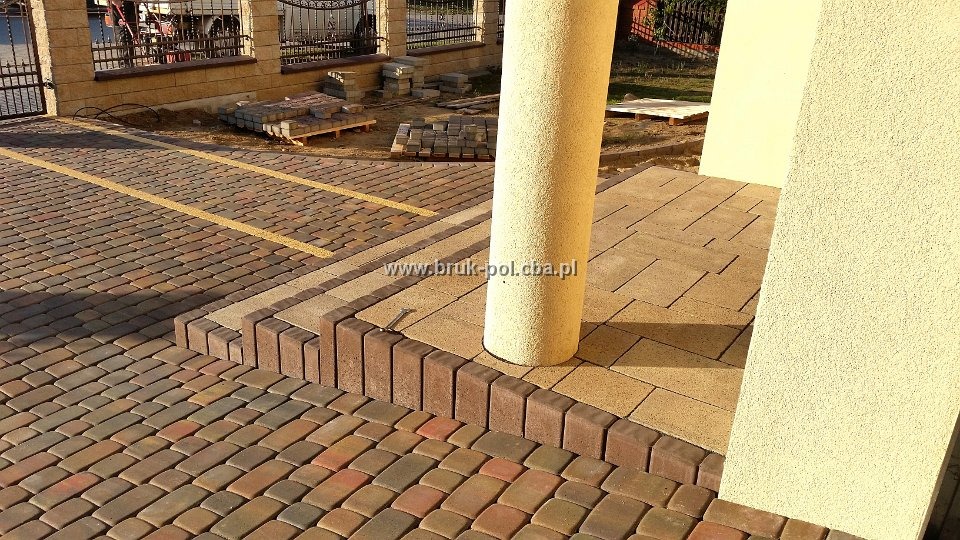 Ułożona kostka brukowa w odcieniach brązu i czerwieni, z żółtymi pasami wyznaczającymi miejsca parkingowe, obrzeża z ciemniejszej kostki, fragment kolumny i ściany budynku w tle.