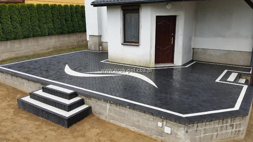 Nowoczesna aranżacja podjazdu z ciemnej kostki brukowej z białymi akcentami, schody wejściowe z kontrastującym wypełnieniem, widoczny fragment domu z brązowymi drzwiami.