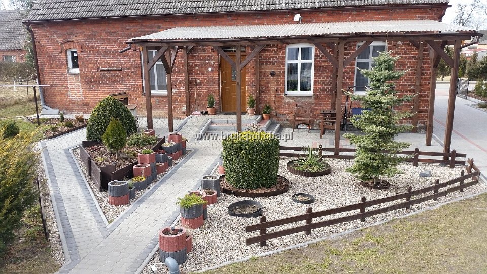 Widok z góry na ogród przed ceglanym domem z drewnianą pergolą, ścieżka z kostki prowadzi przez rabaty i trawnik, widoczne ozdobne donice i niskie ogrodzenie.