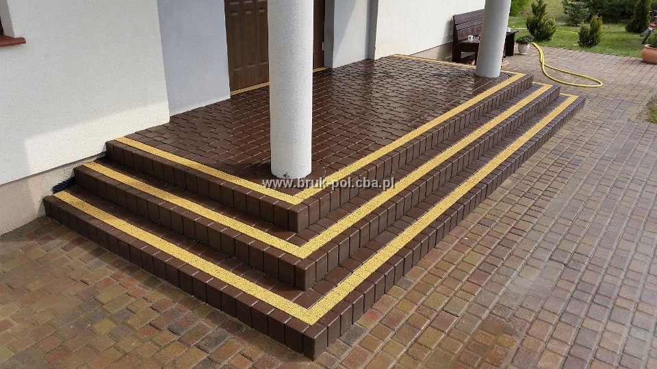 Schody wejściowe do domu wyłożone brązową kostką brukową z żółtymi akcentami, widok z góry, lekko mokra powierzchnia, w tle fragment ogrodu.
