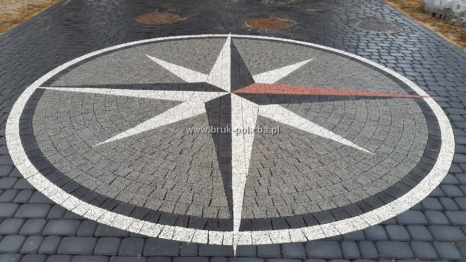 Dekoracyjny wzór kompasu ułożony z kostki brukowej w odcieniach szarości, czerni i bieli, z jednym ramieniem kompasu w kolorze ceglastym, otoczony okręgiem z białej kostki.