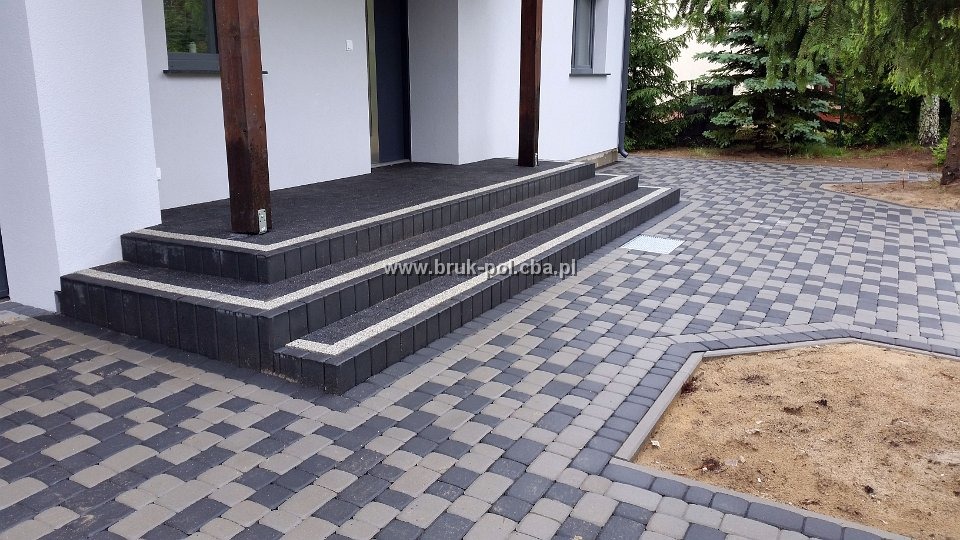 Schody wejściowe do domu wyłożone czarną kostką brukową z jasnymi fugami, płynnie przechodzące w ścieżkę z szaro-czarnej kostki, obrzeża wykończone ciemną kostką, widoczny fragment trawnika i drzew