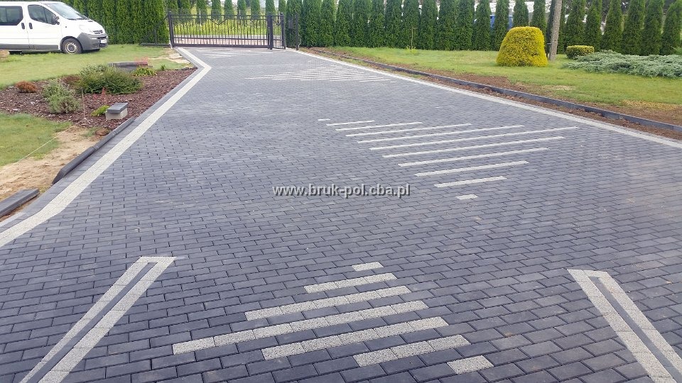 Nowoczesny podjazd z ciemnoszarej kostki brukowej ułożonej we wzór z jasnymi akcentami, prowadzący do bramy wjazdowej i domu z zadbanym ogrodem.