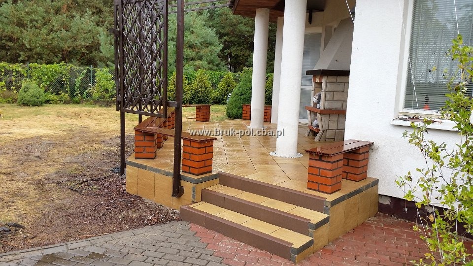 Wyłożony kostką brukową taras z ceglanymi ławkami i schodami, widok na ogród i dom z kolumnami.