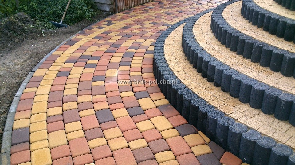 Kolorowa kostka brukowa ułożona w łukowaty wzór, obrzeża schodów z ciemnych betonowych palisad.