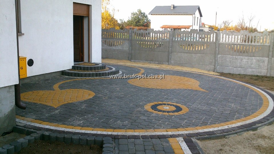 Okrągły podjazd z kostki brukowej w kolorach szarości i żółci, z dekoracyjnymi wzorami w kształcie serca i koła; widoczne schody wejściowe do domu, skrzynka gazowa i betonowe ogrodzenie.