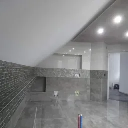 Nowoczesna łazienka na poddaszu z szarymi płytkami, mozaiką i podświetlanym lustrem, widoczne rury instalacyjne wystające z podłogi.