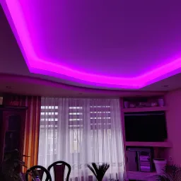 Salon z podwieszanym sufitem oświetlonym fioletowym światłem LED, meblami w stylu retro i oknem z białymi firankami w tle.