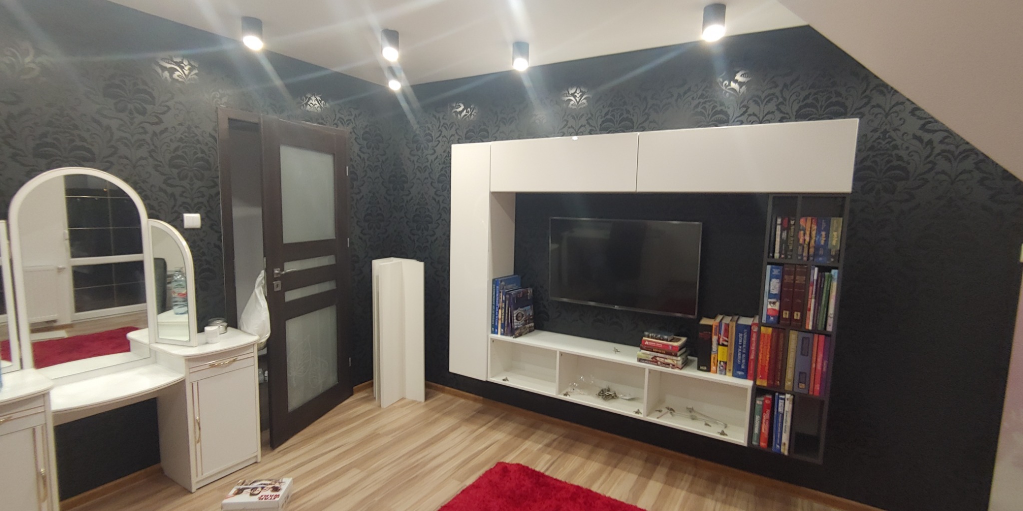 Eleganckie wnętrze pokoju z czarną tapetą w kwiaty, białymi meblami, telewizorem i książkami, oświetlone punktowymi lampami sufitowymi.