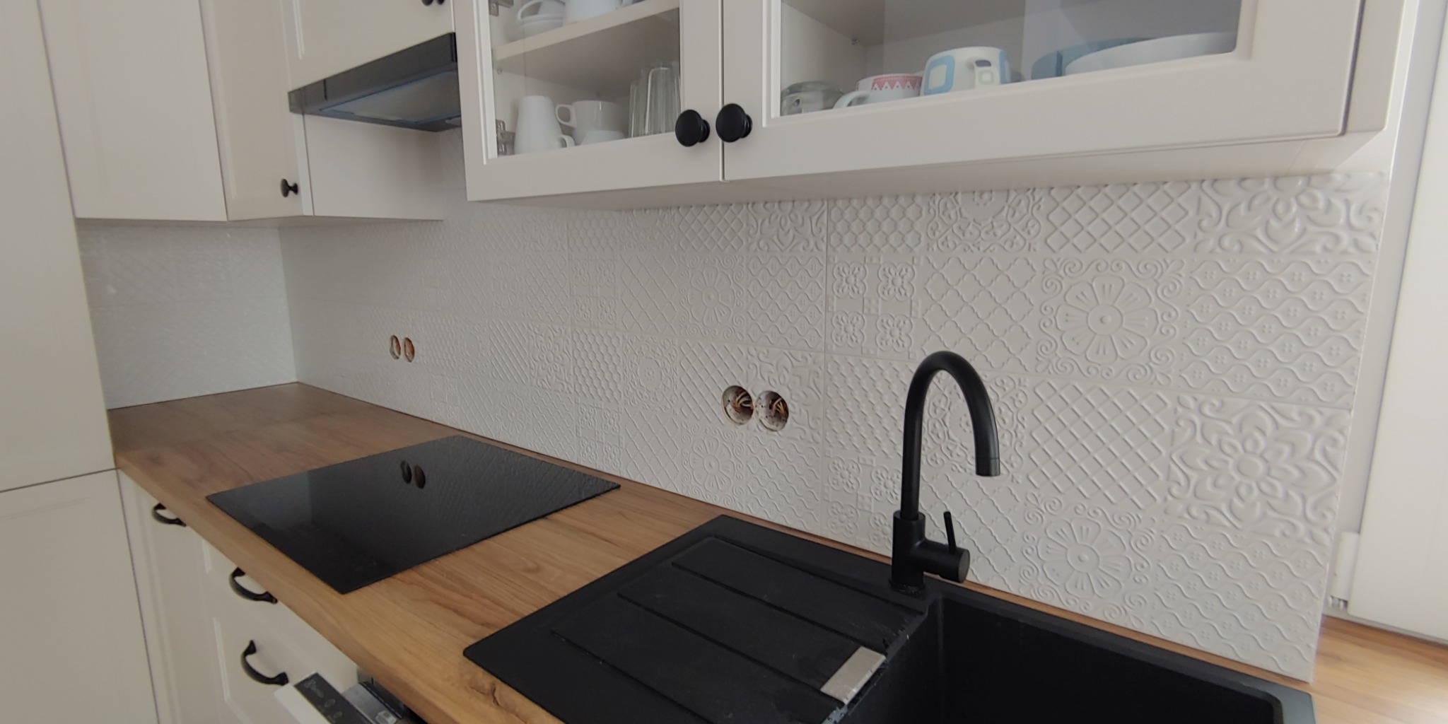 Biała kuchnia z drewnianym blatem, czarnym zlewem i płytą indukcyjną. Na ścianie położone białe, wzorzyste płytki ceramiczne z widocznymi otworami na gniazdka elektryczne.