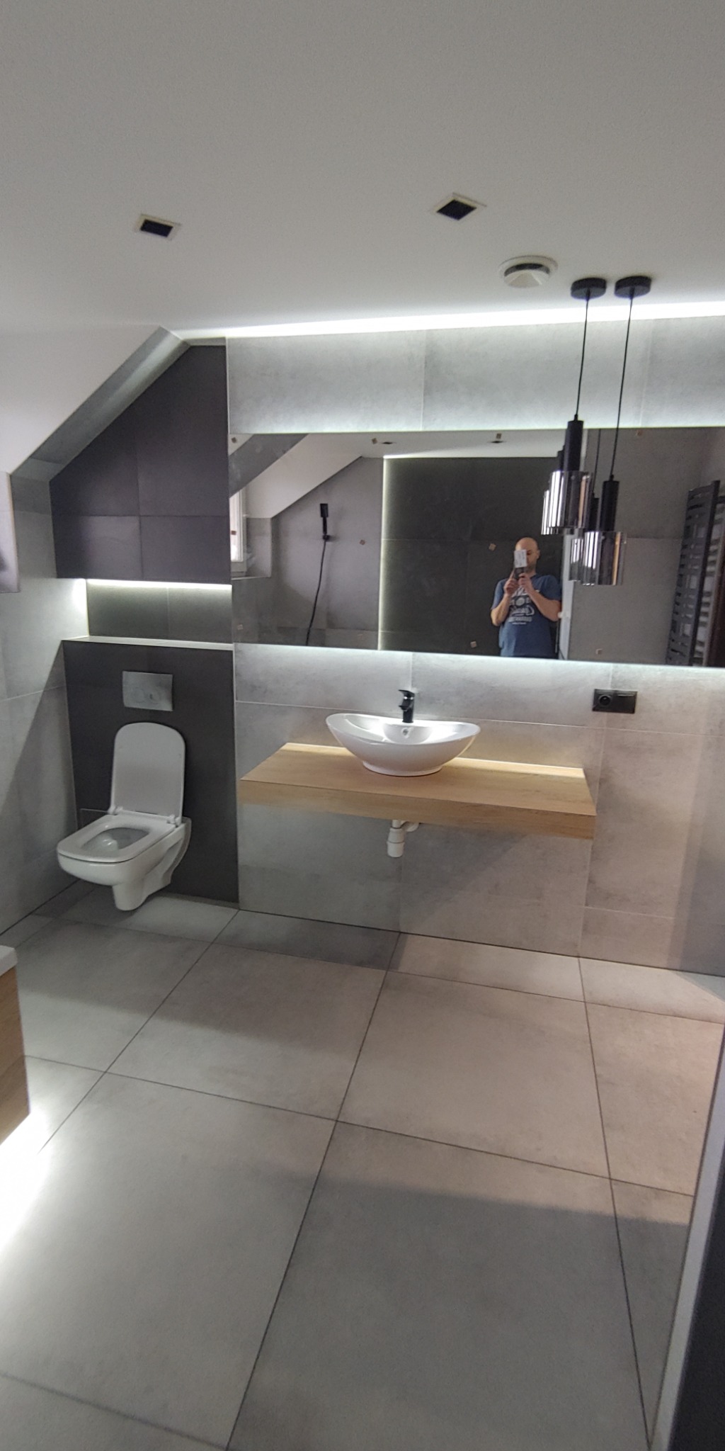 Nowoczesna łazienka w szarościach z umywalką nablatową na drewnianym blacie, oświetleniem LED za lustrem i minimalistyczną toaletą pod skosem dachu.