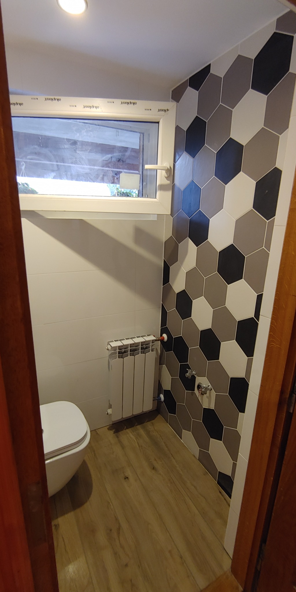 Ukończona łazienka z heksagonalnymi płytkami w odcieniach szarości i czerni na jednej ścianie, białe płytki na pozostałych ścianach, drewnopodobna podłoga, biały grzejnik i toaleta pod oknem.