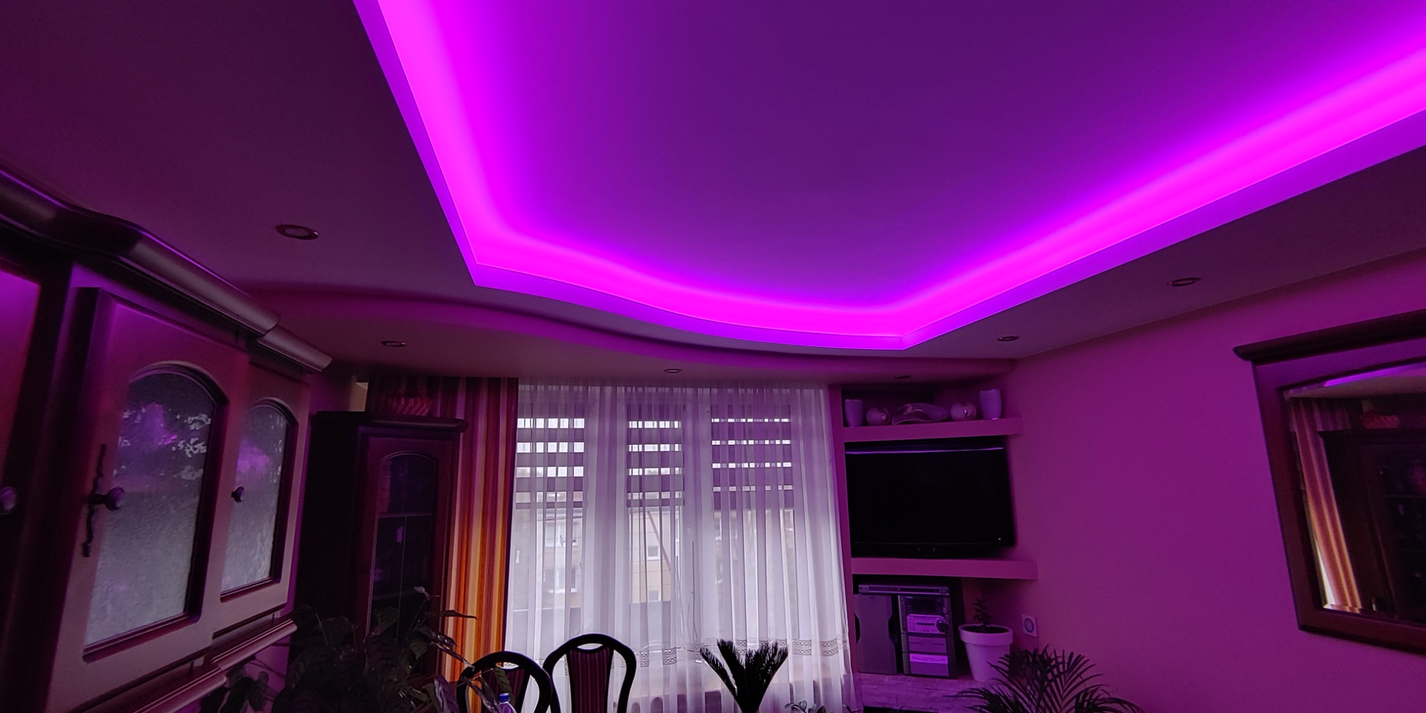 Salon z podwieszanym sufitem oświetlonym fioletowym światłem LED, meblami w stylu retro i oknem z białymi firankami w tle.