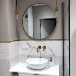 Nowoczesna łazienka z okrągłym lustrem, złotymi akcentami i umywalką nablatową, podwieszane żarówki edison nad umywalką, marmurowe płytki na ścianie.