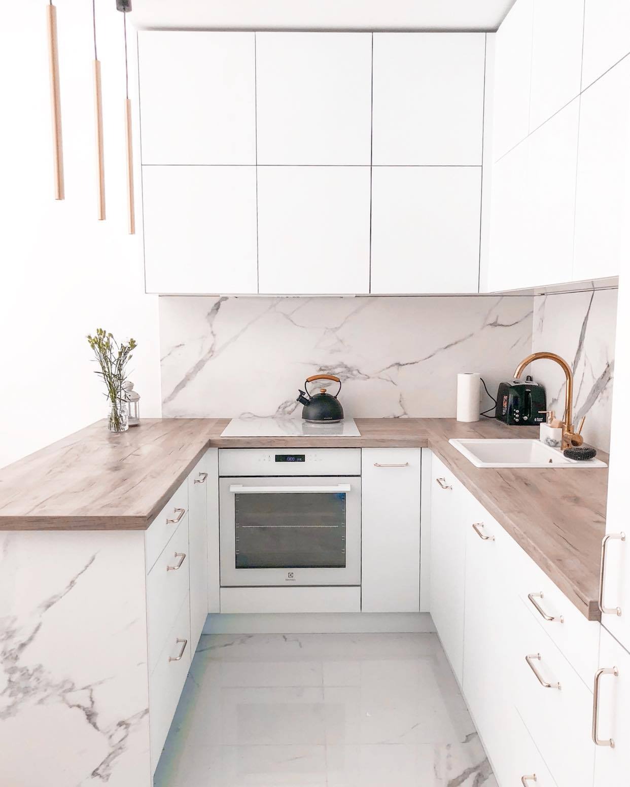 Jasna kuchnia w kształcie litery U, z białymi szafkami z marmurowym wzorem, drewnianym blatem i złotą baterią, czajnik na płycie indukcyjnej.