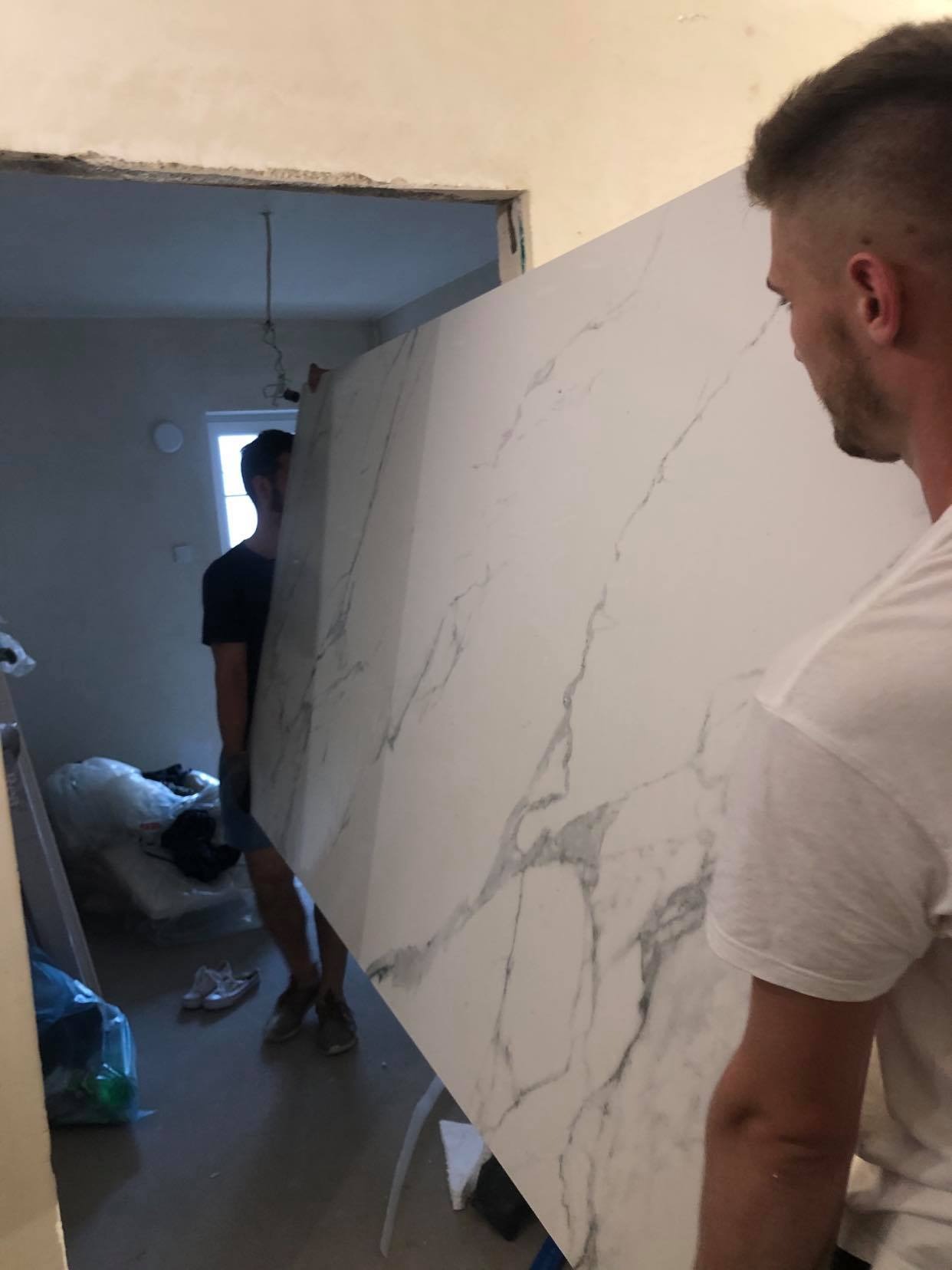 Dwóch mężczyzn przenosi dużą płytę z białego marmuru do remontowanego pomieszczenia z widoczną instalacją elektryczną na suficie i oknem w tle.