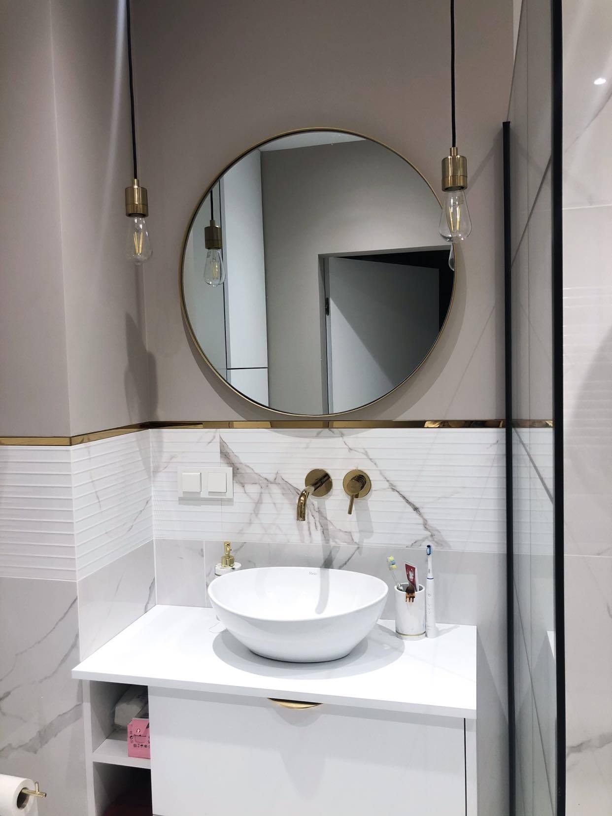 Nowoczesna łazienka z okrągłym lustrem, złotymi akcentami i umywalką nablatową, podwieszane żarówki edison nad umywalką, marmurowe płytki na ścianie.