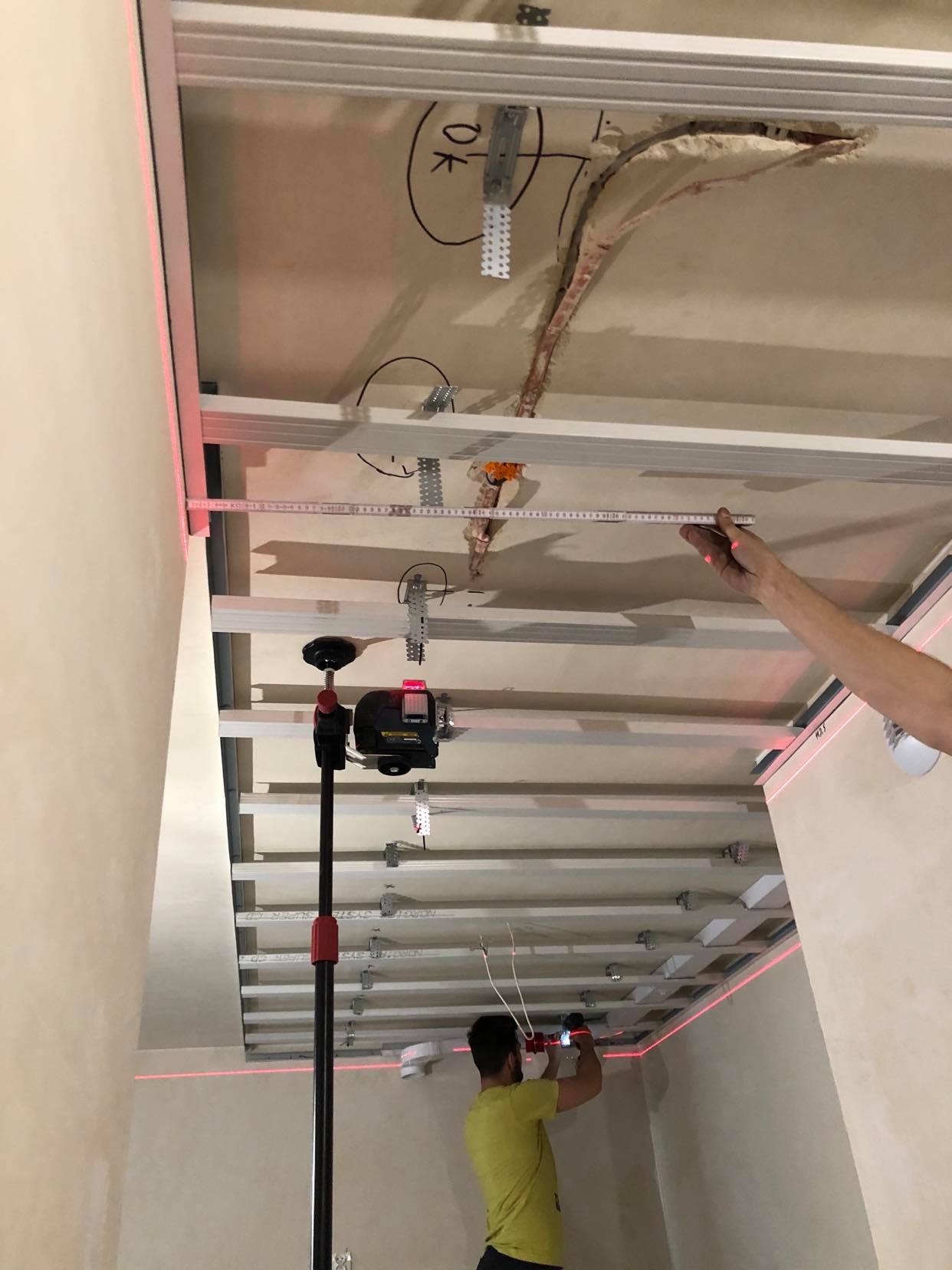 Montaż stelaża sufitu podwieszanego z płyt kartonowo-gipsowych, widoczny laser krzyżowy i pomiar odległości, instalacja elektryczna w suficie, napis 'OK' na starym suficie.