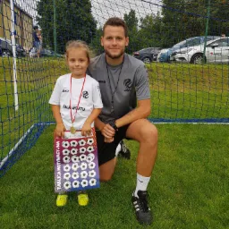 Dziewczynka w koszulce 'FALA SPORTU' z medalem, trzymająca tablicę motywacyjną z piłkami nożnymi, stoi obok trenera na tle siatki bramki i zielonej trawy.