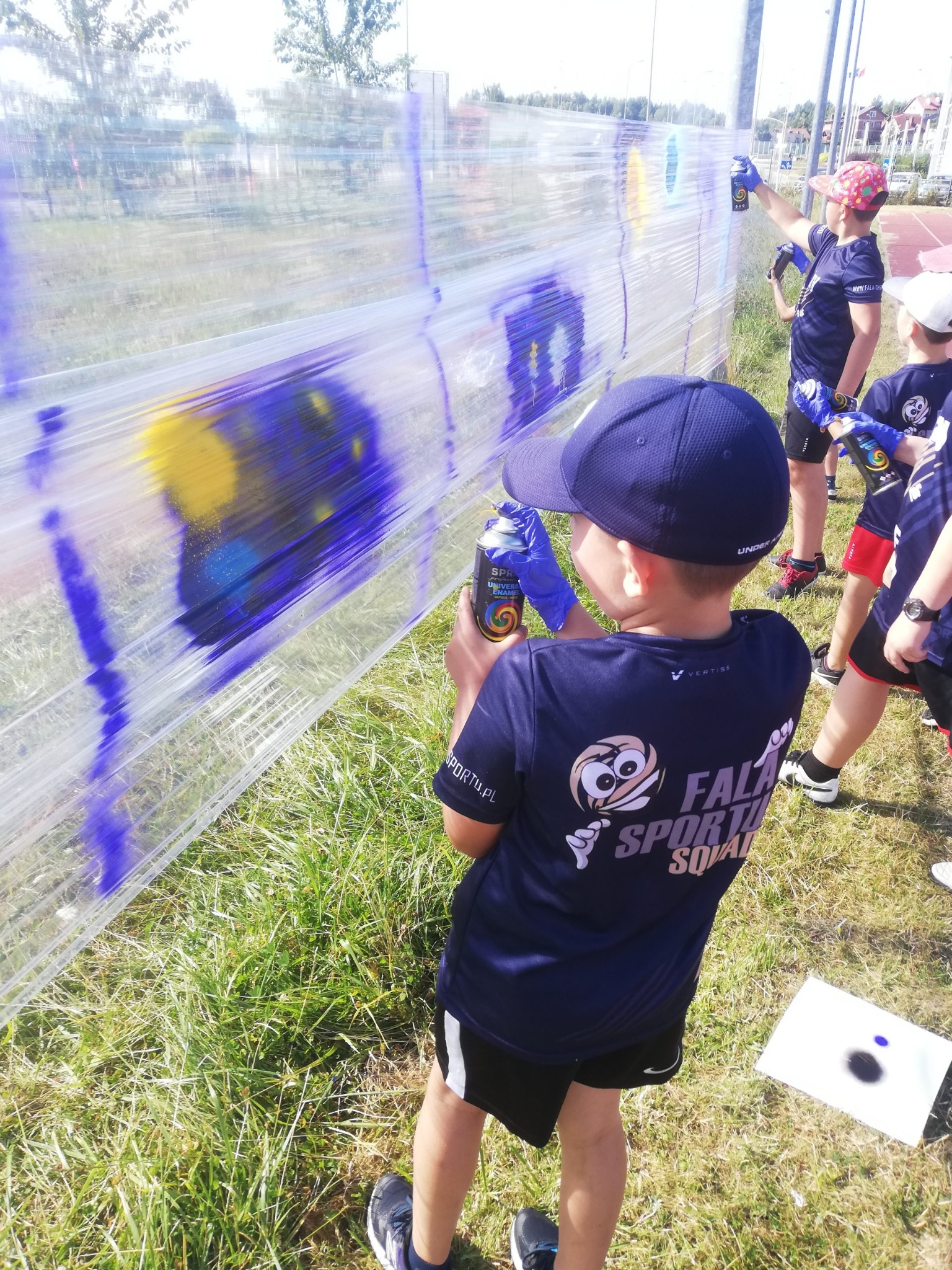 Grupa dzieci w niebieskich koszulkach maluje sprayami na przezroczystej folii rozpiętej na zewnątrz, tworząc kolorowe abstrakcje.