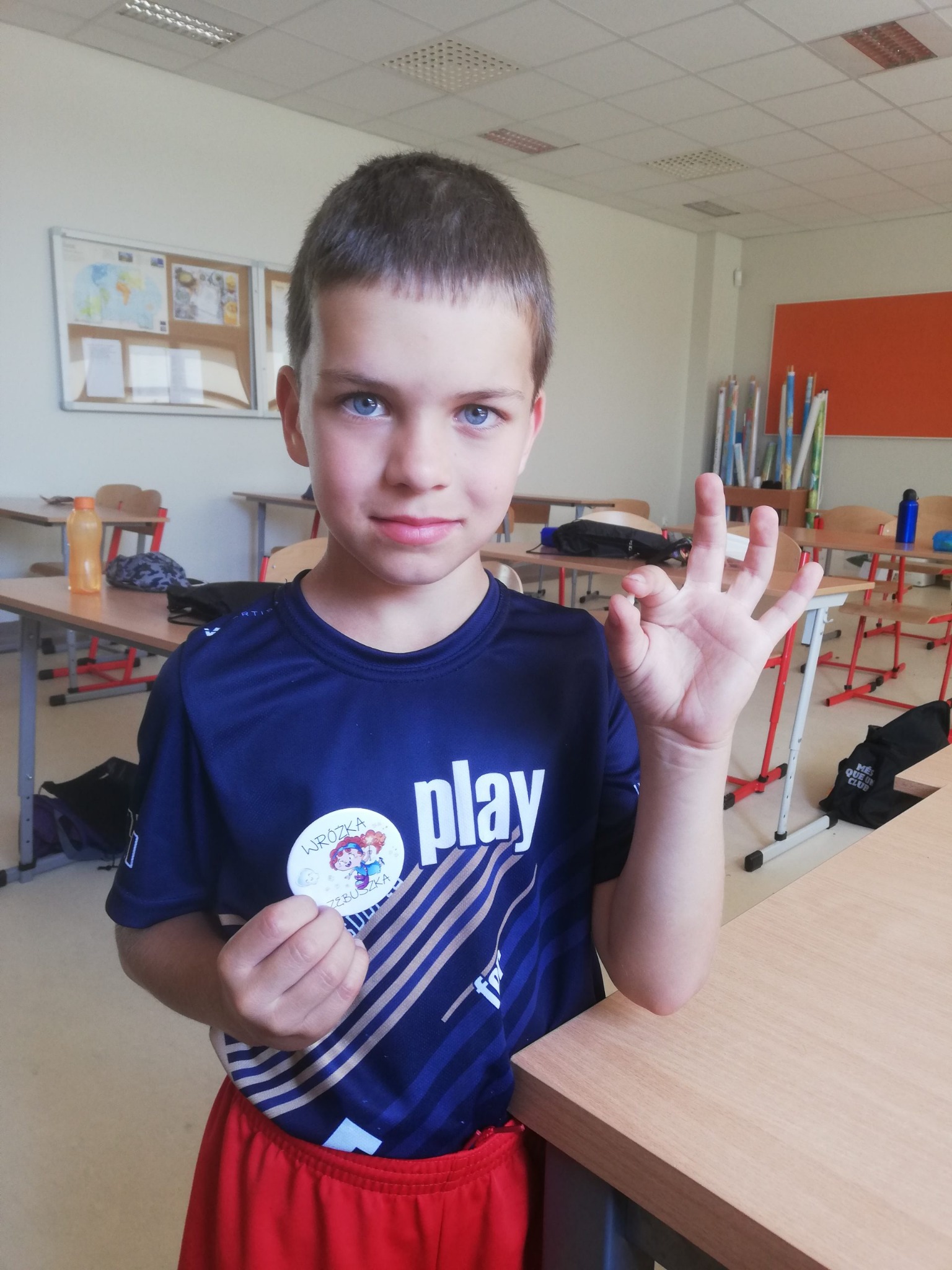 Chłopiec w koszulce z napisem 'play' i przypinką 'Wróżka Zębuszka' w klasie pokazuje znak OK.