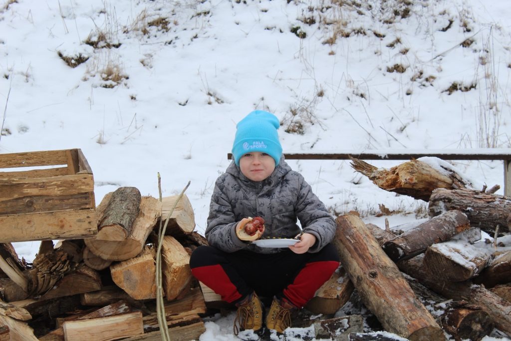 Chłopiec w niebieskiej czapce 'Fala Sportu' siedzi w Gdańsku na tle zaśnieżonego krajobrazu i drewna, trzymając talerz z hot dogiem.