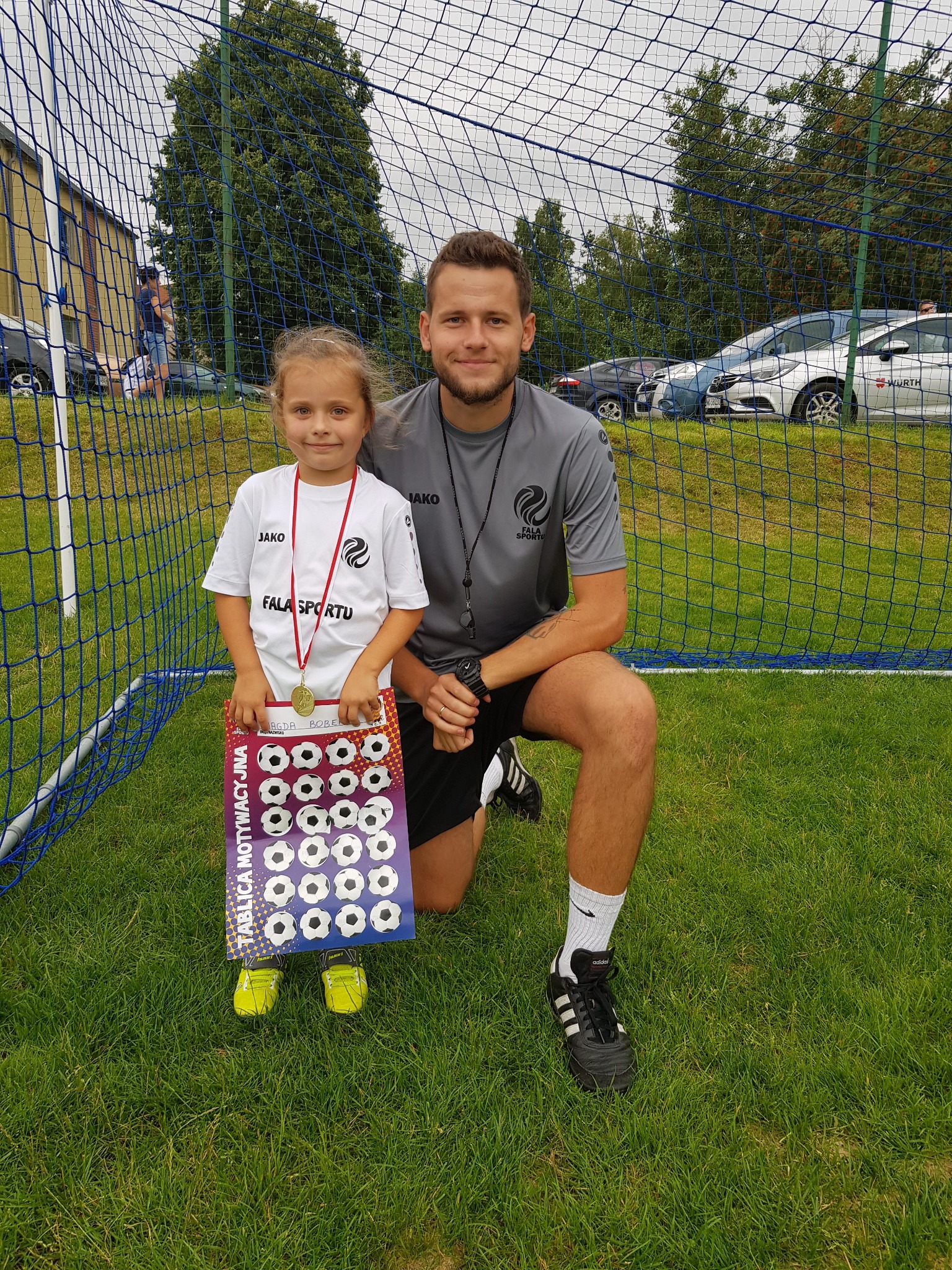 Dziewczynka w koszulce 'FALA SPORTU' z medalem, trzymająca tablicę motywacyjną z piłkami nożnymi, stoi obok trenera na tle siatki bramki i zielonej trawy.