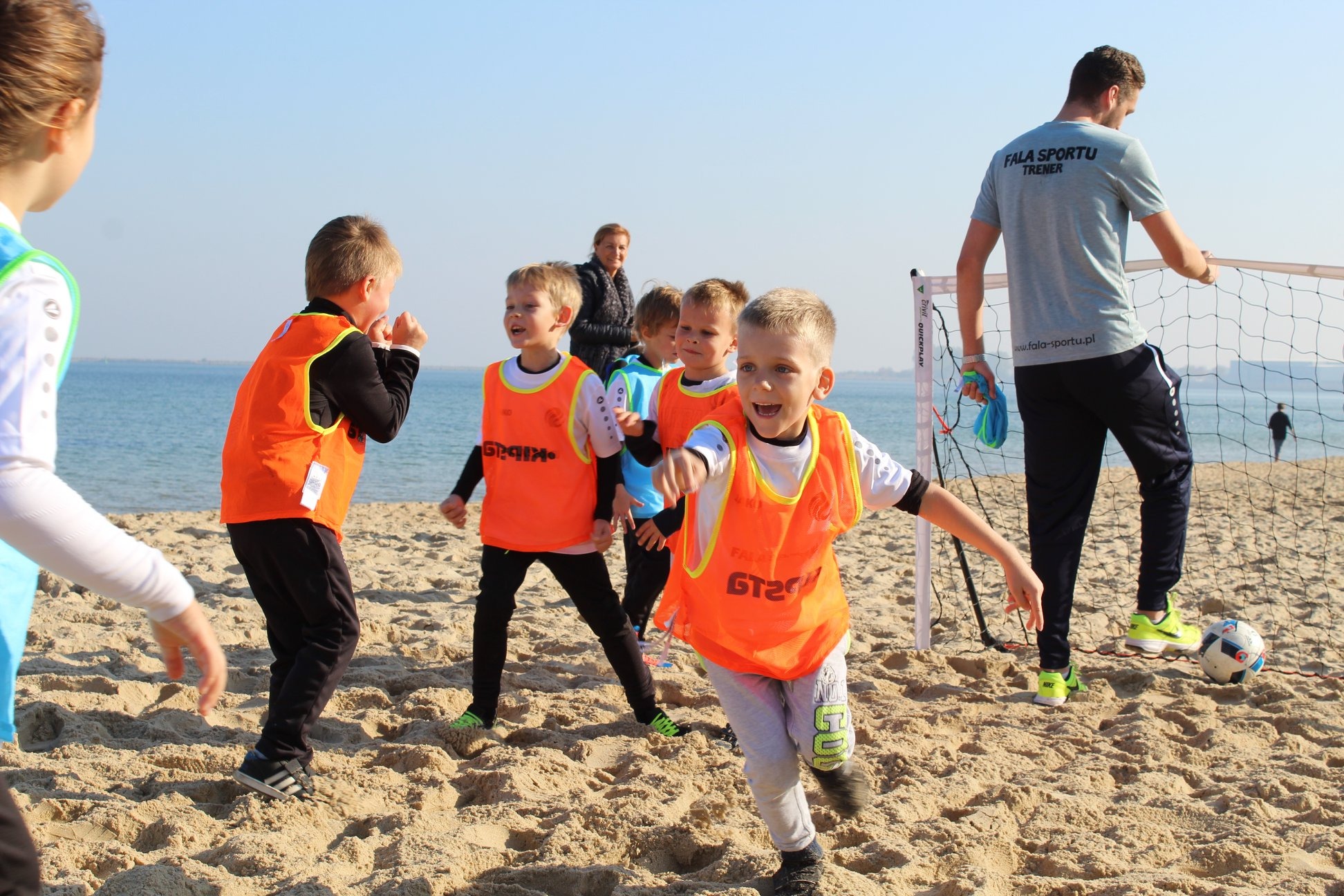 Trening piłkarski dla dzieci na piaszczystej plaży z widokiem na morze, chłopiec w pomarańczowej kamizelce z żółtymi obrzeżami biegnie w kierunku obiektywu, a w tle stoi trener przy siatce.