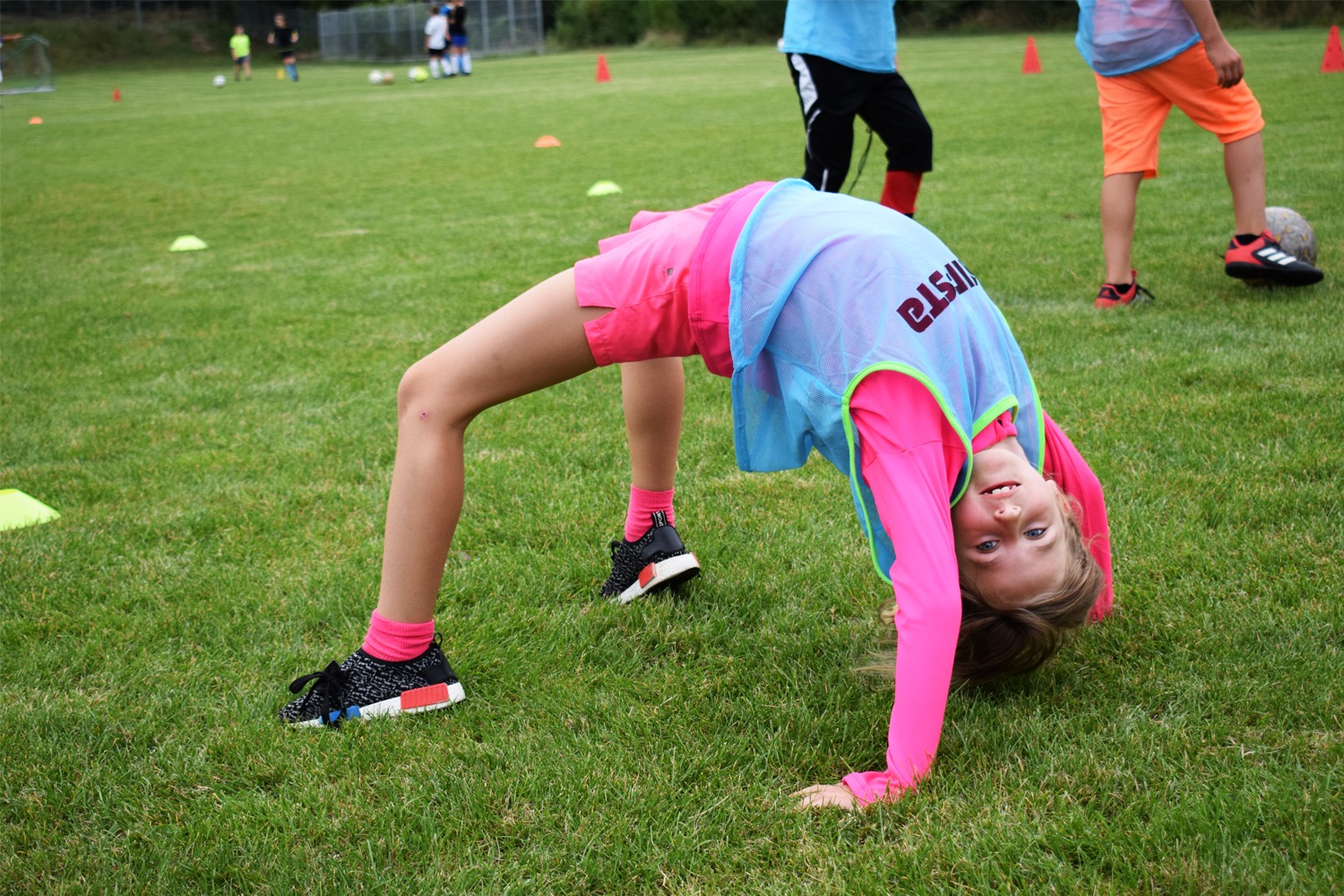 Dziewczynka w stroju sportowym, robiąca mostek na zielonej trawie podczas treningu piłkarskiego, w tle widoczni inni uczestnicy i piłki.