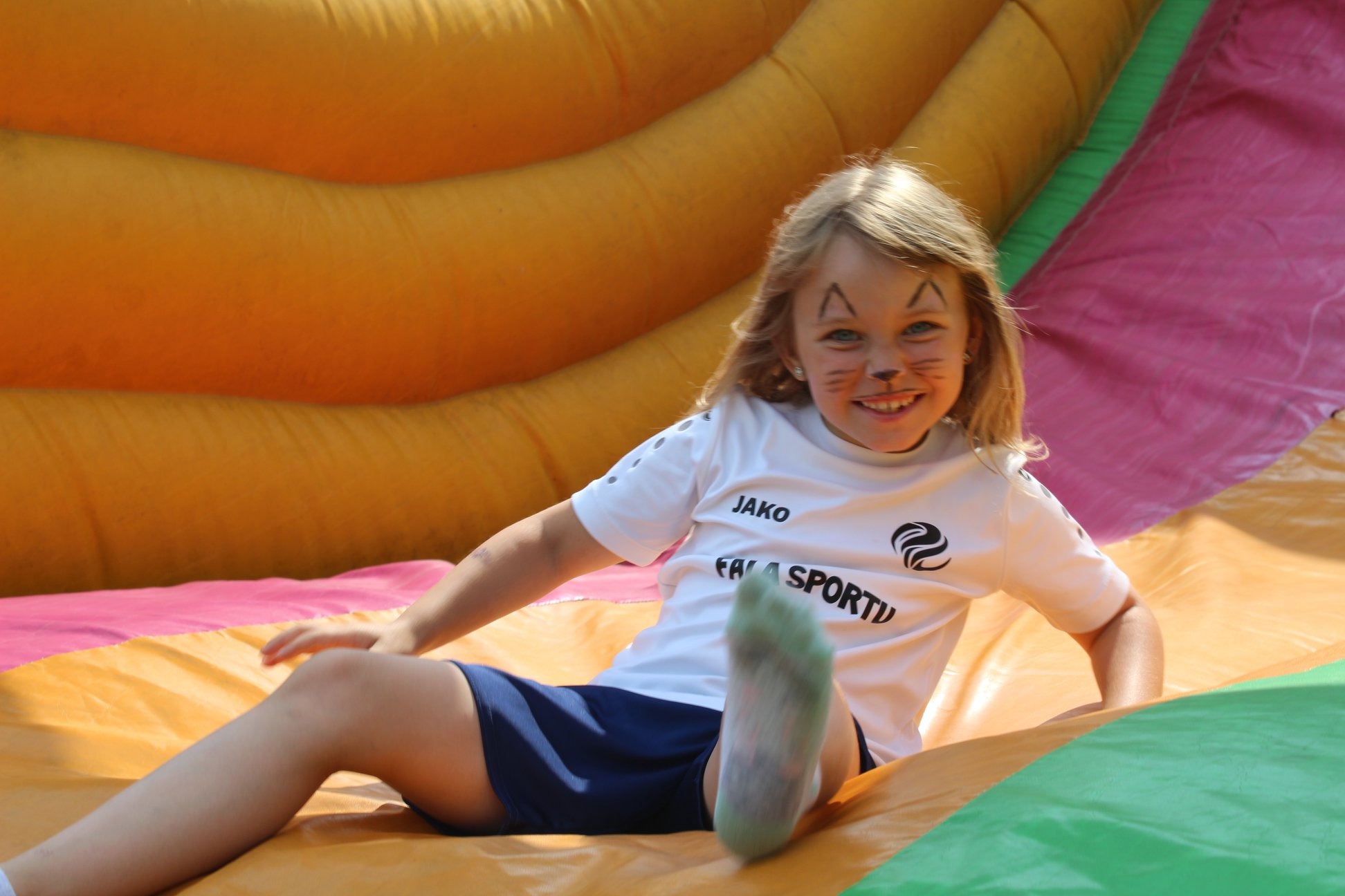 Uśmiechnięta dziewczynka z pomalowaną twarzą leży na dmuchanym zamku, ubrana w sportowy strój. Widoczne elementy zamku w kolorach żółtym, różowym i zielonym.