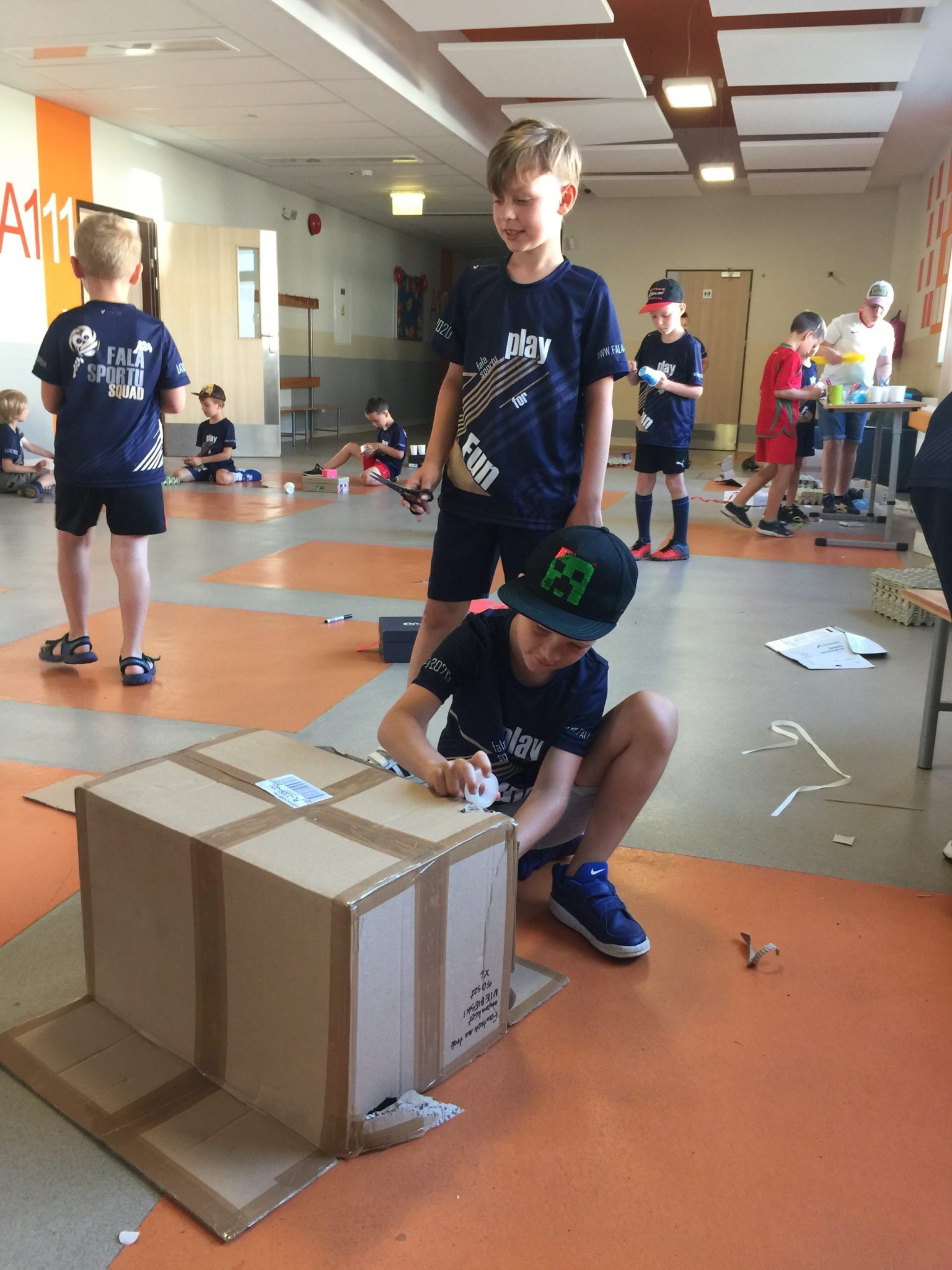 Grupa dzieci w koszulkach z nadrukiem w jasnym pomieszczeniu, tworząca konstrukcje z kartonowych pudeł, używając taśmy klejącej i nożyczek.