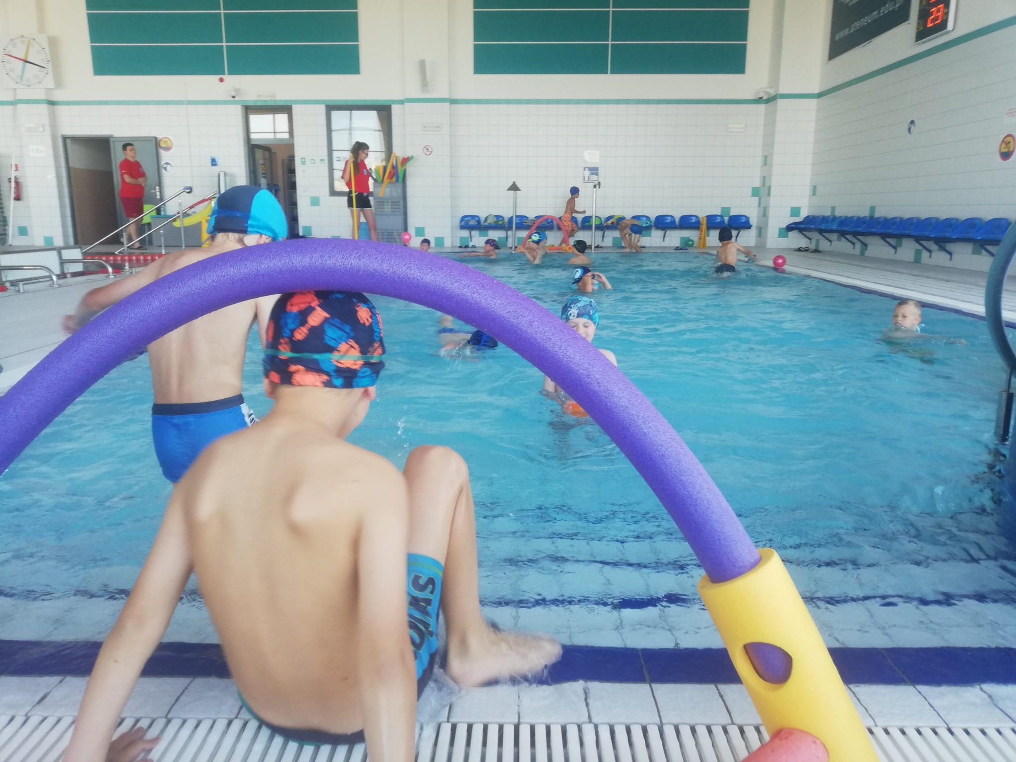 Dzieci w czepkach i strojach kąpielowych bawią się w basenie krytym, widoczna niebieska woda i kolorowe makarony do pływania, na brzegu siedzi chłopiec z mokrymi stopami.