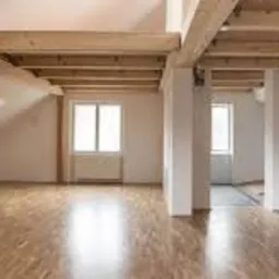 Jasne, przestronne wnętrze poddasza z drewnianymi belkami stropowymi i świeżo ułożoną, błyszczącą podłogą parkietową, widok na okno i drzwi w tle.