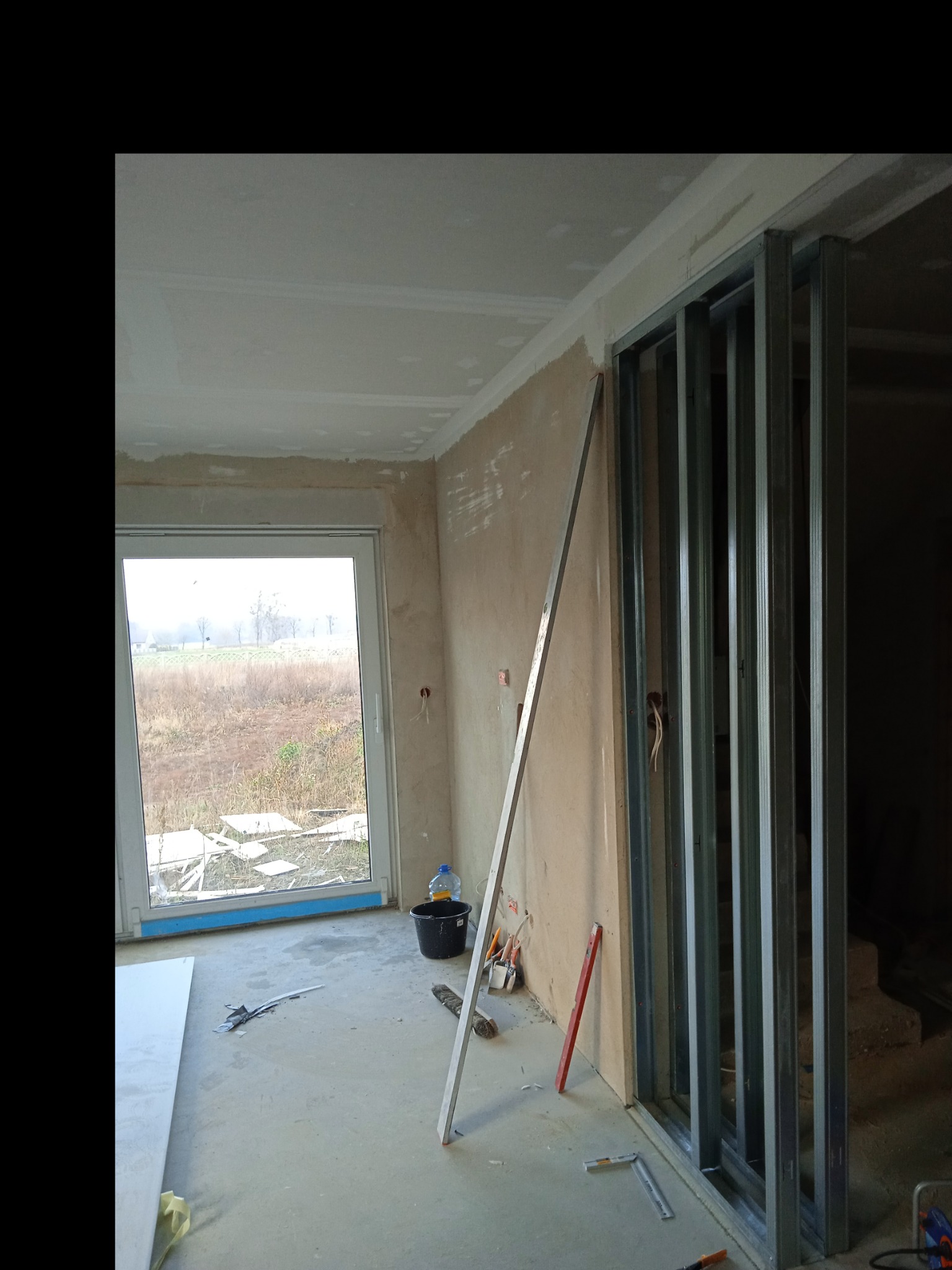 Wnętrze w trakcie prac wykończeniowych: widoczne metalowe profile ścianki działowej, częściowo wykończona gładź na ścianach i suficie, okno z widokiem na zewnątrz, narzędzia budowlane rozłożone...