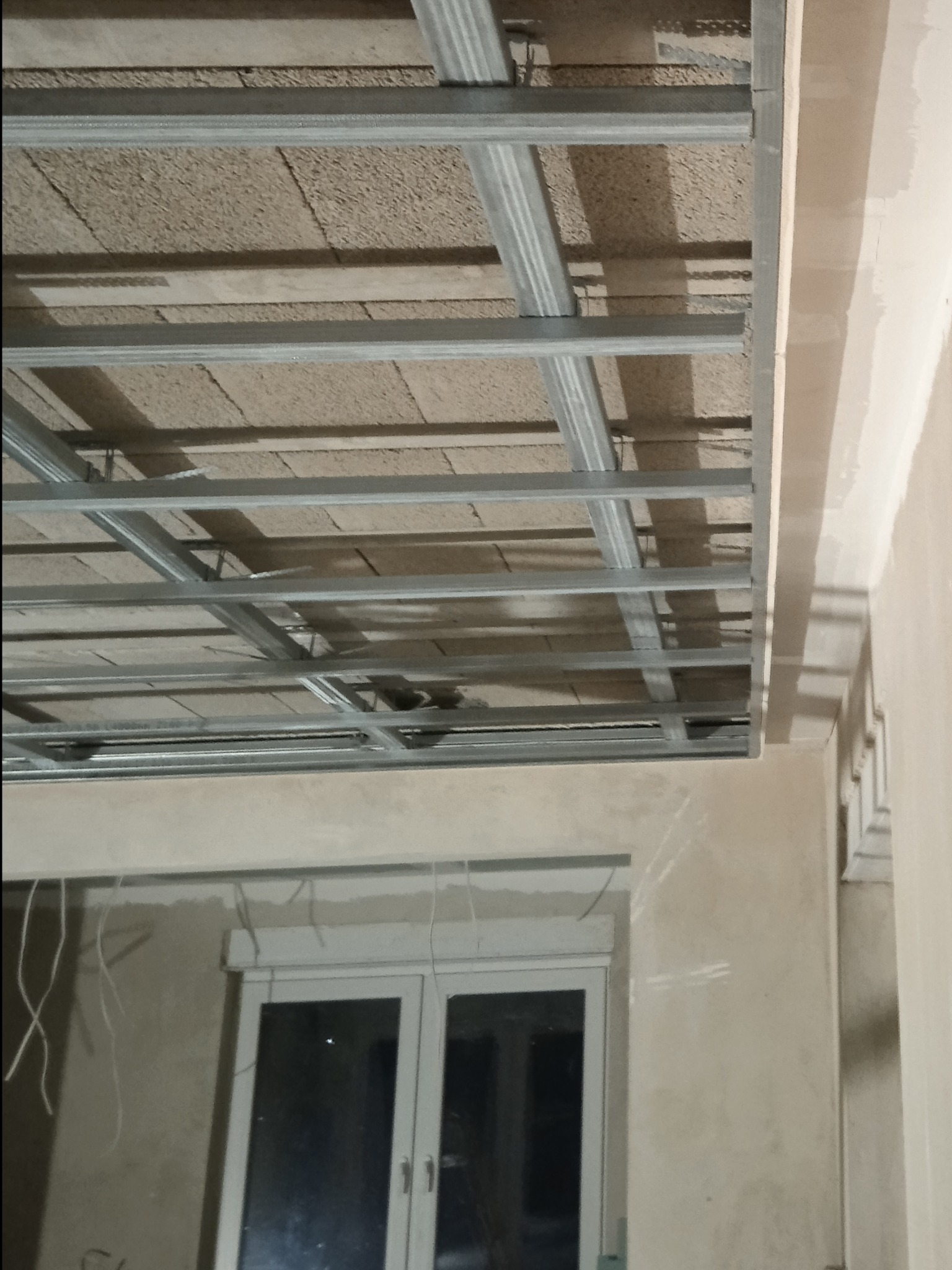 Metalowa konstrukcja sufitu podwieszanego z płytami OSB w trakcie montażu, widok z dołu, surowe ściany w tle, okno z roletą.