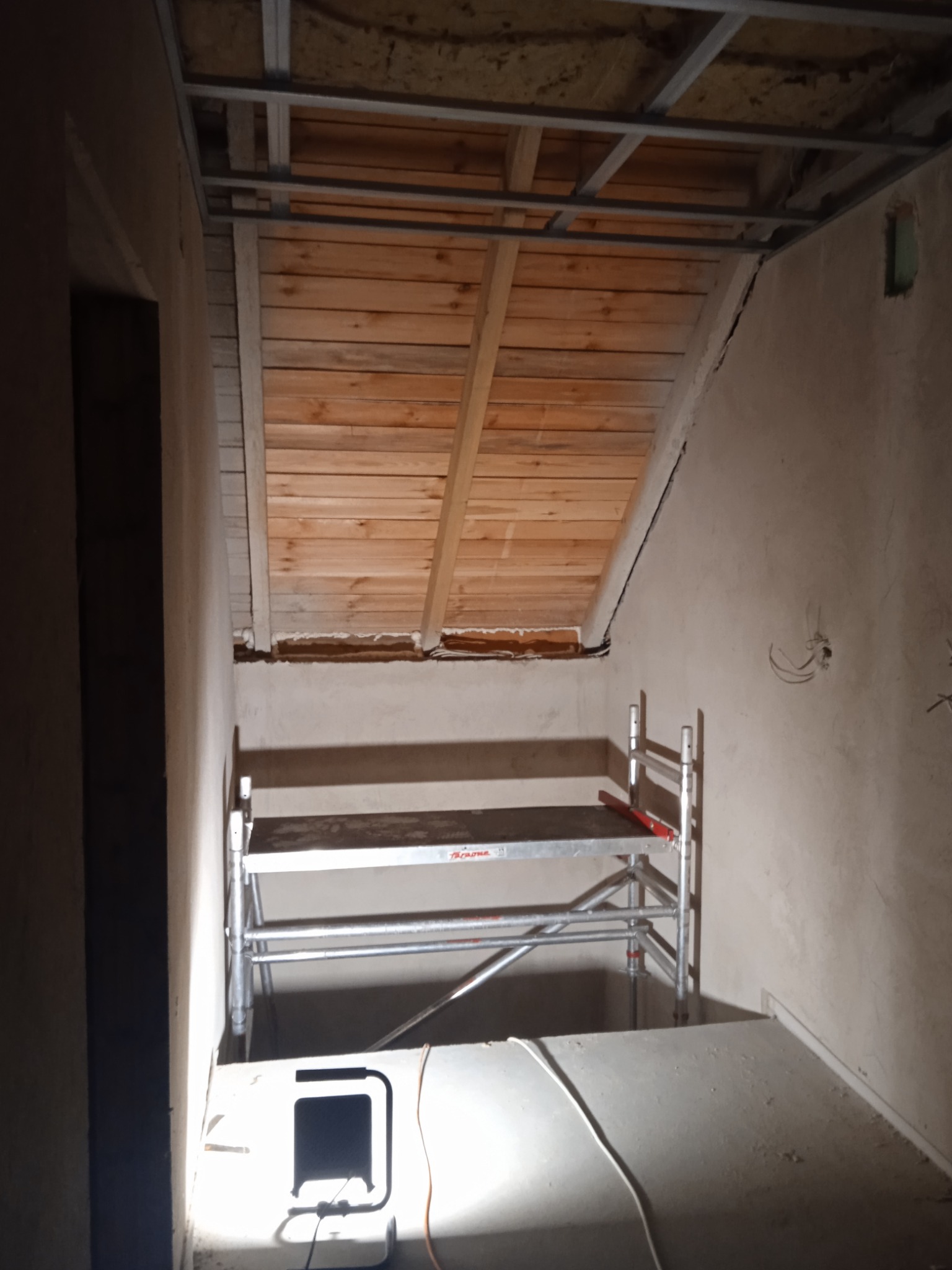 Wnętrze w trakcie remontu: surowe ściany, drewniane deski poddasza, metalowy stelaż pod sufitem, aluminiowe rusztowanie i lampa halogenowa oświetlająca podłogę.