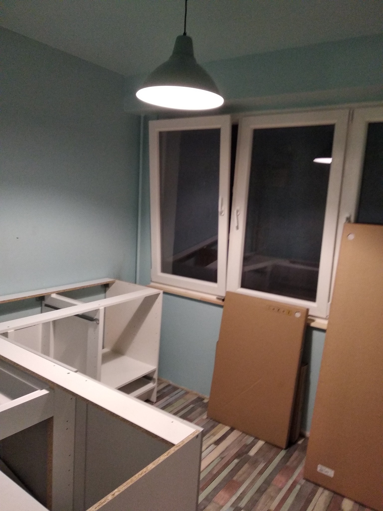 Montaż białych szafek kuchennych w surowym stanie, widoczne elementy konstrukcyjne i płyty meblowe oparte o ścianę przy oknie.