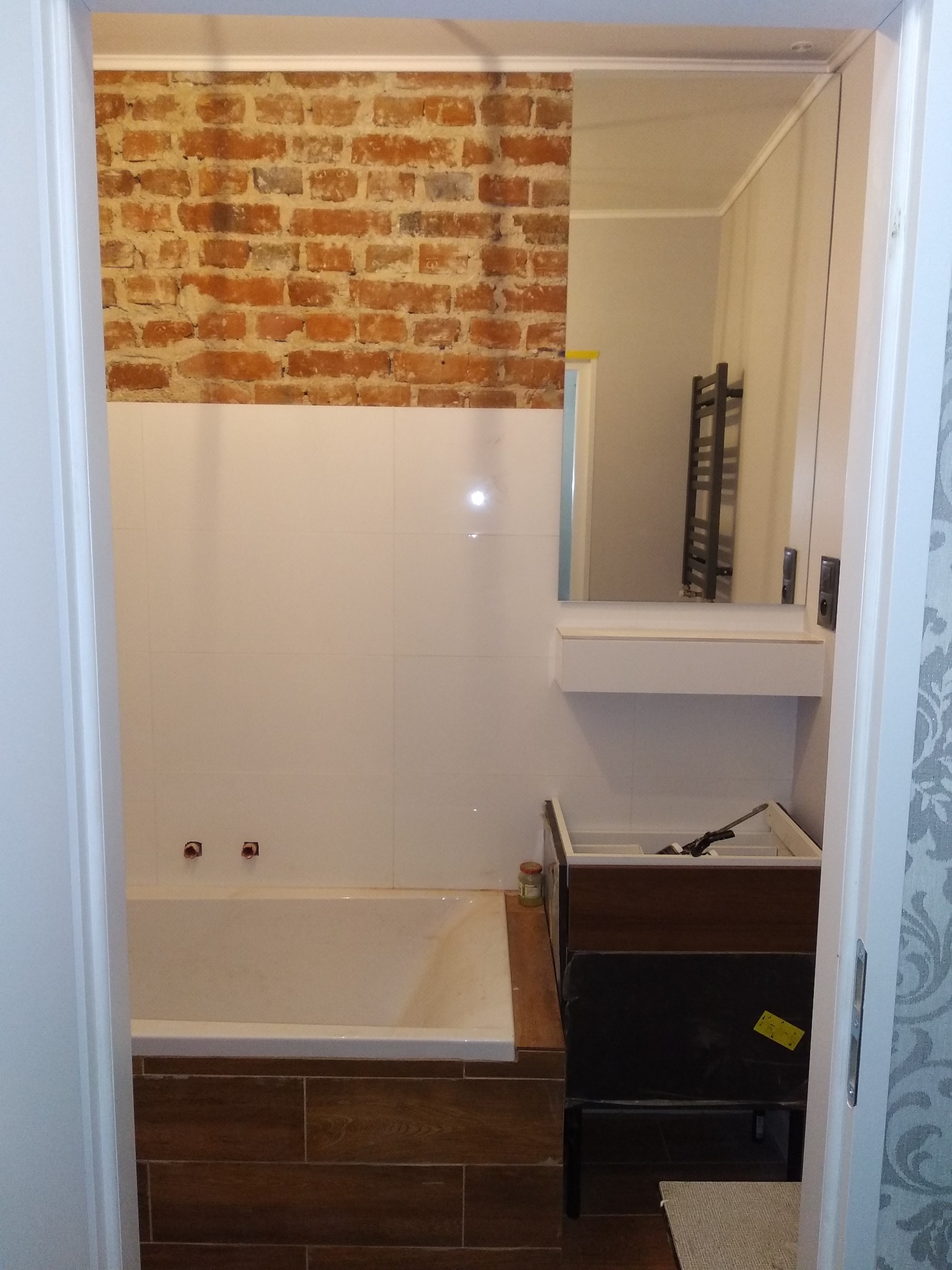 Wnętrze łazienki w trakcie remontu, widoczna wanna z obudową imitującą drewno, białe płytki ścienne, fragment ściany z cegły, niedokończona szafka pod umywalkę z narzędziami w środku i lustro...