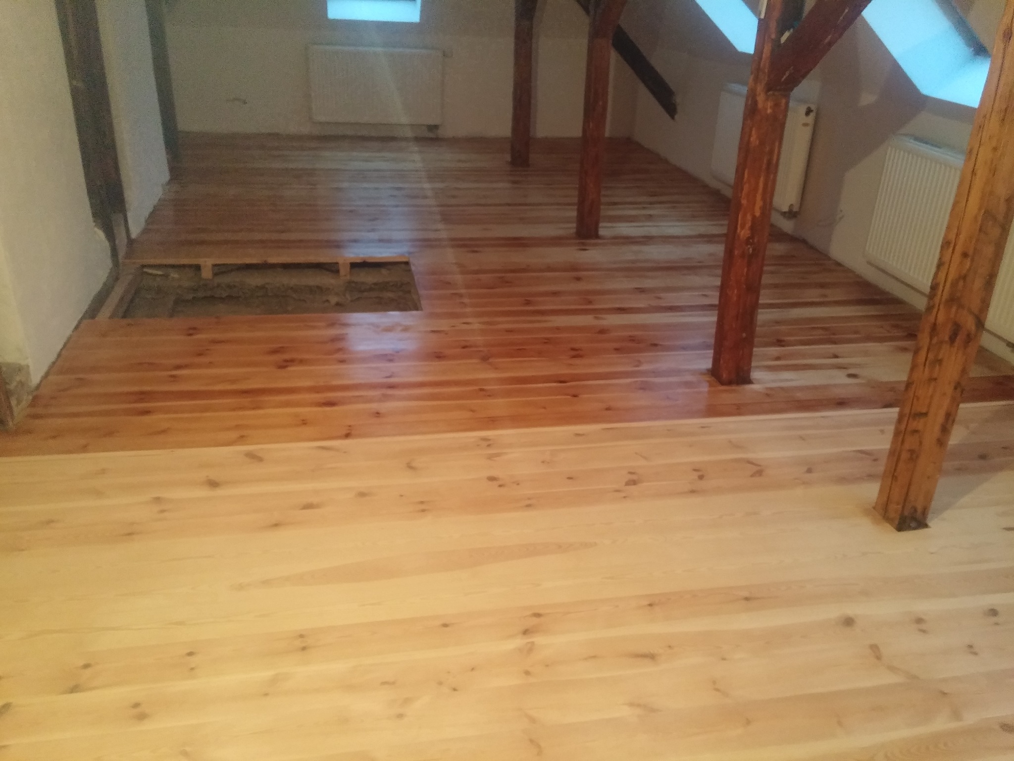 Fragment poddasza z drewnianą podłogą w trakcie układania, widoczna różnica w odcieniach drewna, częściowo wykończona podłoga i otwór w podłodze z widocznym podłożem.