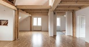 Jasne, przestronne wnętrze poddasza z drewnianymi belkami stropowymi i świeżo ułożoną, błyszczącą podłogą parkietową, widok na okno i drzwi w tle.