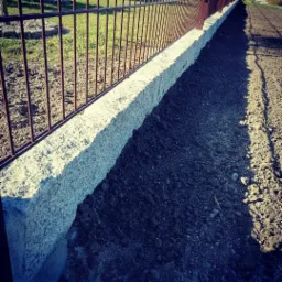 Granitowa obrzeże trawnika z metalowym ogrodzeniem, świeżo przygotowana ziemia pod nasadzenia, widok z bliska.