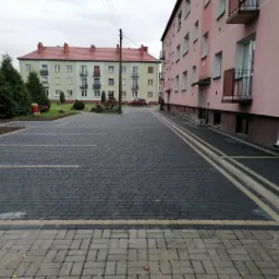 Nowo ułożona, ciemnoszara kostka brukowa na parkingu osiedlowym z wyznaczonymi miejscami postojowymi, obrzeża z jaśniejszej kostki, budynki mieszkalne w tle.