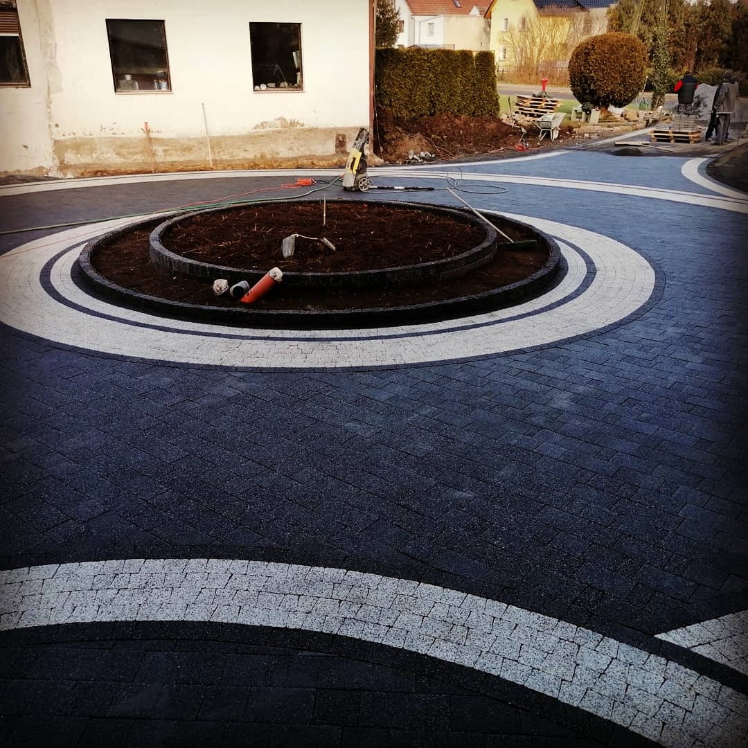 Okrągły podjazd z ciemnej kostki brukowej z białymi akcentami i okrągłym klombem pośrodku w trakcie realizacji, widoczne narzędzia i węże, w tle budynek i drzewa.