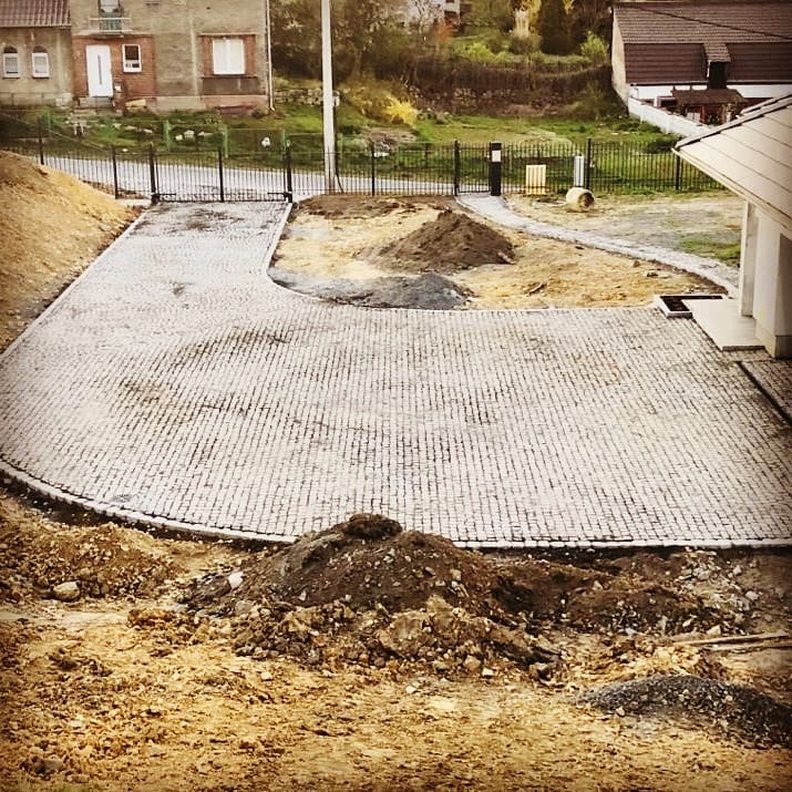 Ułożona szara kostka na podjeździe, widoczne przygotowane podłoże i ogrodzenie w tle.