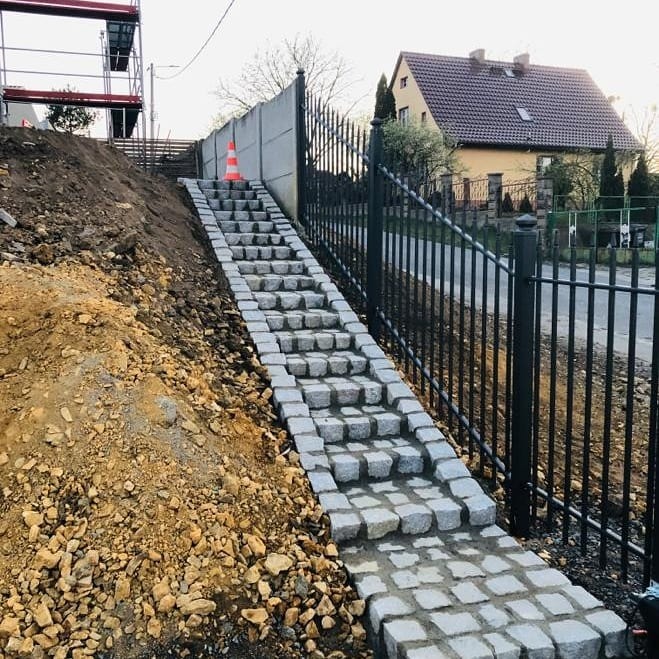 Schody z kostki granitowej prowadzące po zboczu, z widocznym ogrodzeniem i domem w tle.