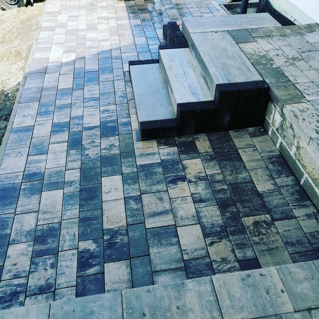 Ułożona nawierzchnia z kostki brukowej w odcieniach szarości i błękitu, z widocznymi schodami wykończonymi czarnymi obrzeżami, tworząca estetyczny wygląd podwórka.