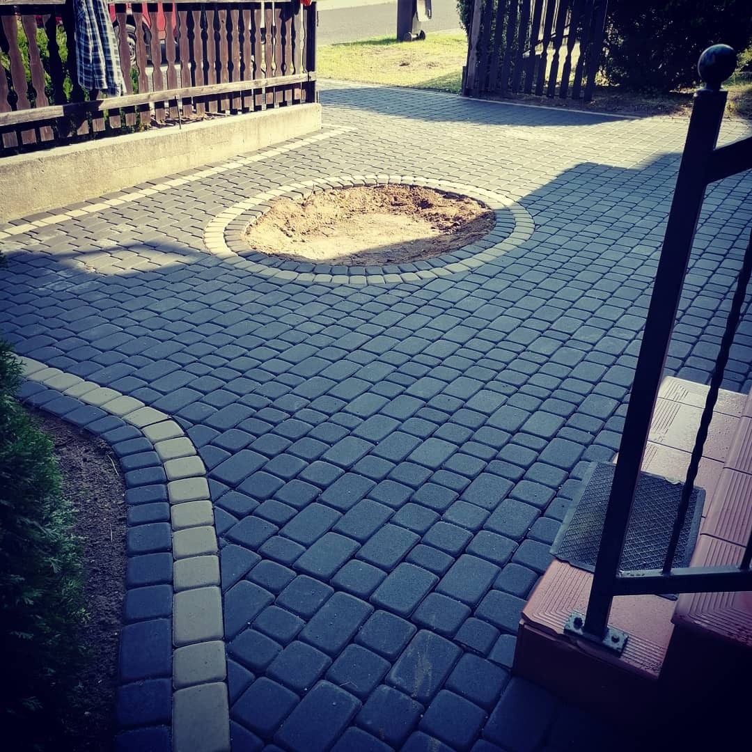 Świeżo ułożona, ciemnoszara kostka brukowa z obramowaniem z jaśniejszych kostek, tworząca okrągłe zagłębienie wypełnione piaskiem, widok z perspektywy schodów.