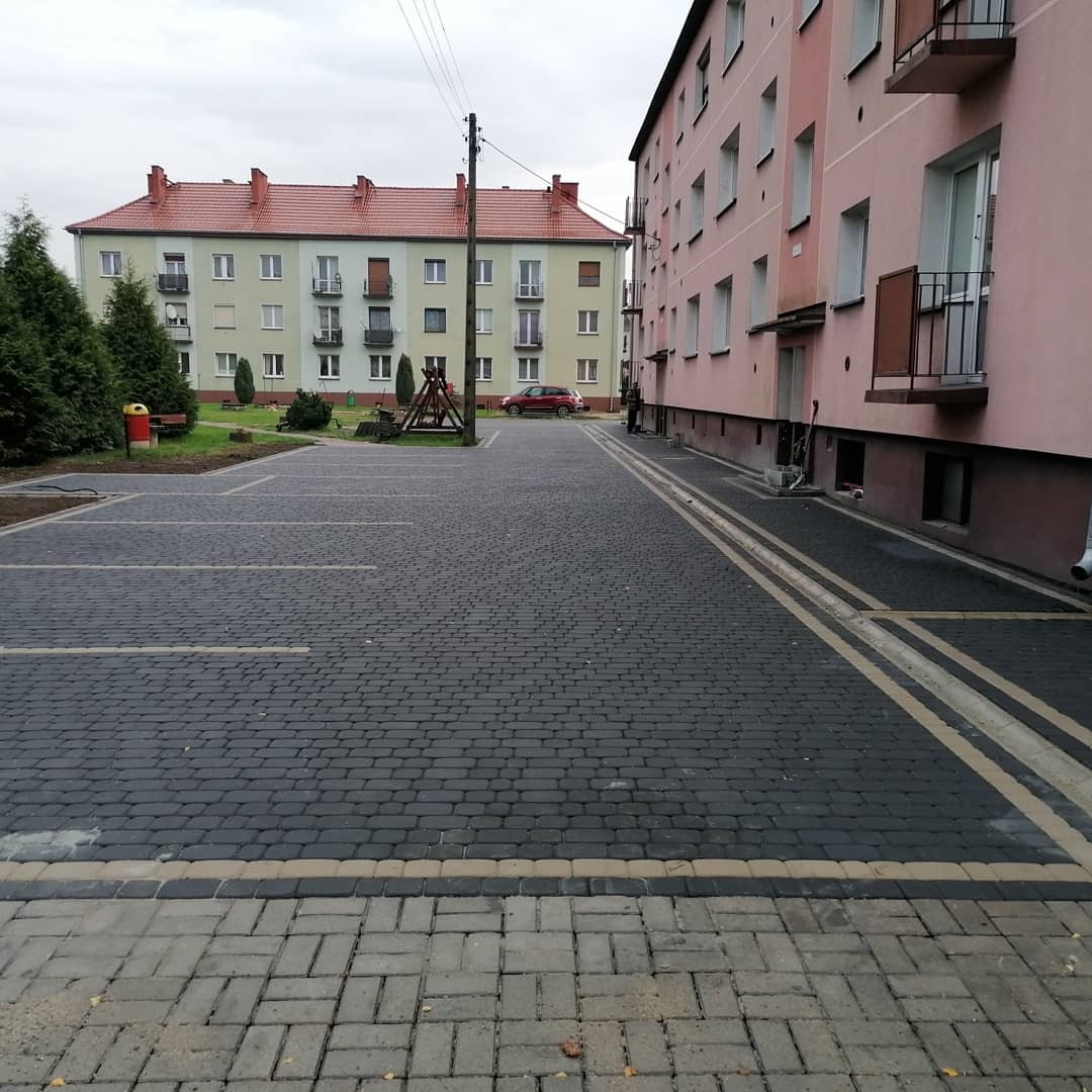 Nowo ułożona, ciemnoszara kostka brukowa na parkingu osiedlowym z wyznaczonymi miejscami postojowymi, obrzeża z jaśniejszej kostki, budynki mieszkalne w tle.